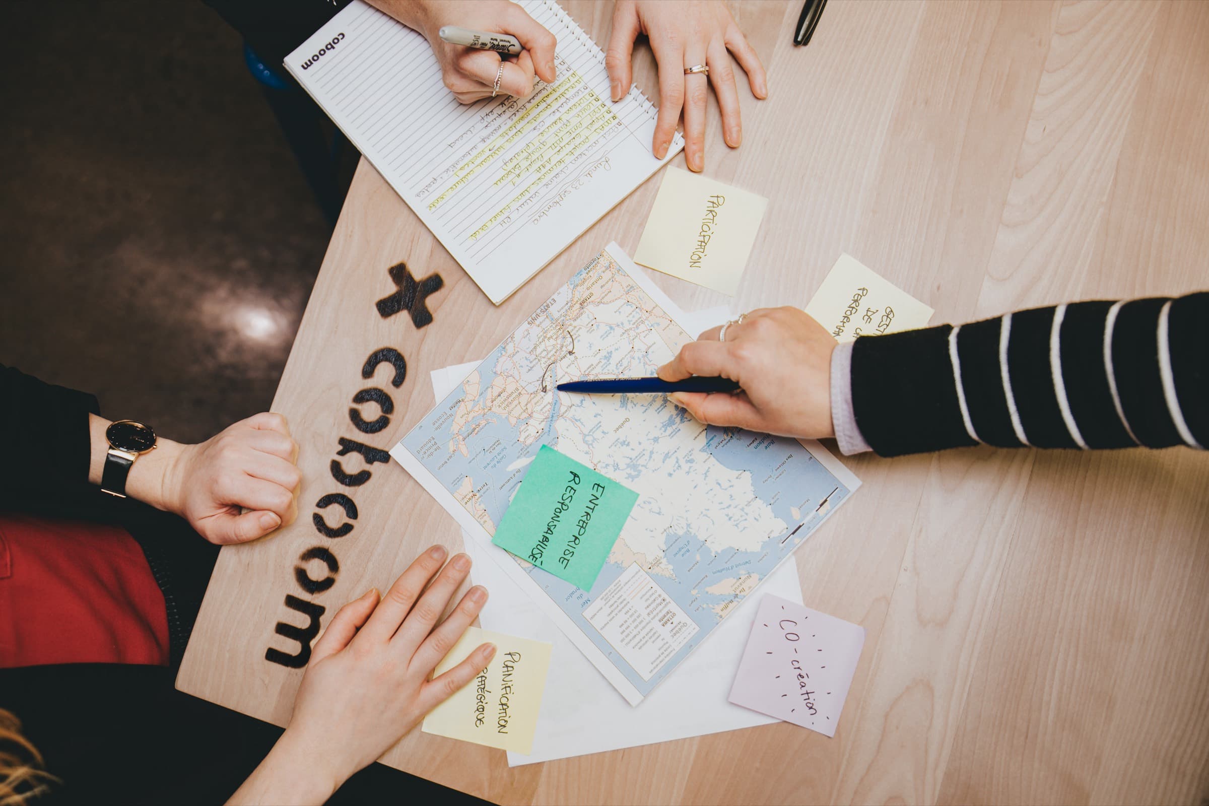 Vue du dessus d'une table d'atelier Coboom : trois personnes annotent une carte du Québec entourée de papillons adhésifs et de carnets.