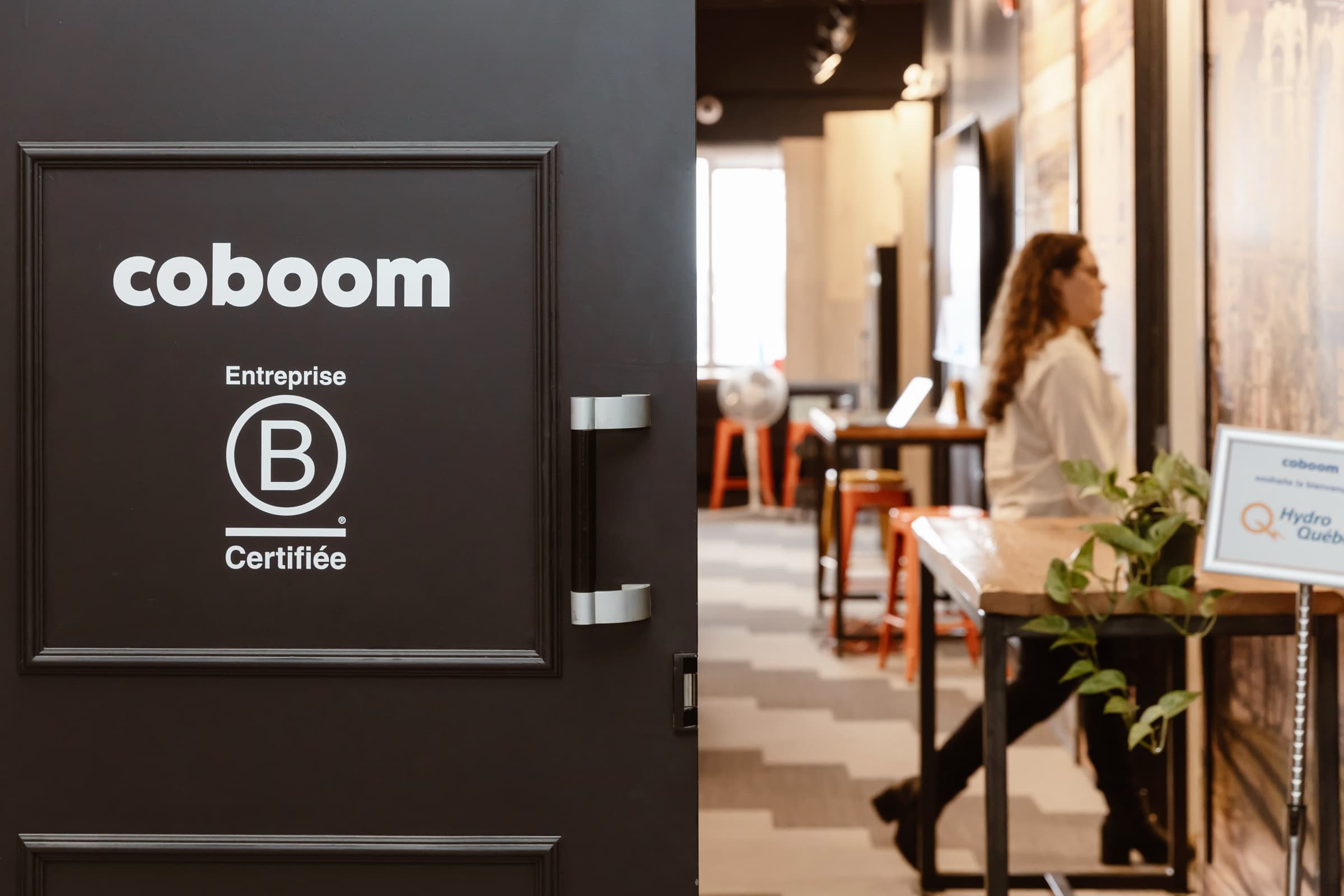 Porte noire du bureau de Coboom : le nom et le sceau « Entreprise B Certifiée » en blanc. Par l'entrebâillement, une échappée sur l'espace de travail.