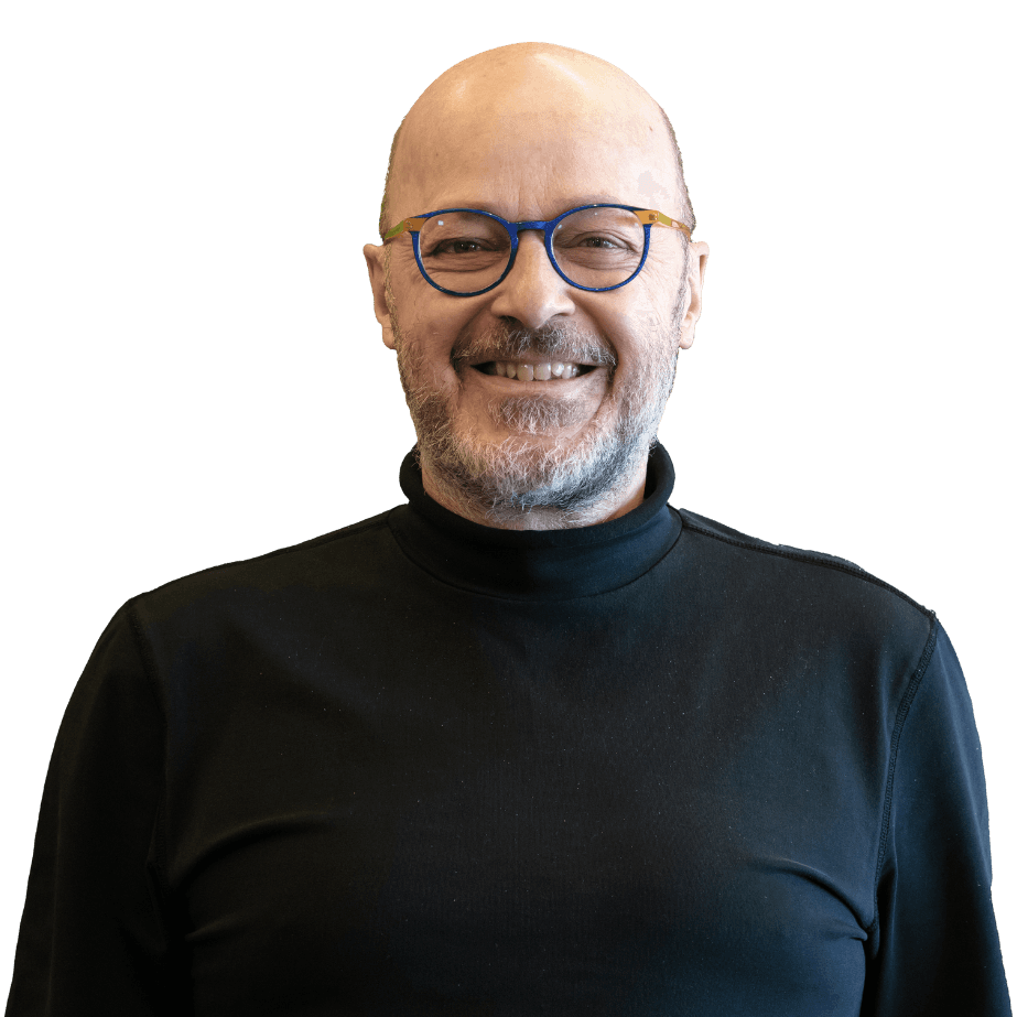 Jean-Pierre Dubé, souriant, crâne dégarni, barbe grise courte, lunettes rondes à monture bleue, col roulé noir.